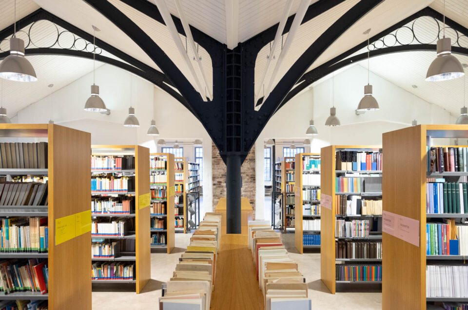 Wide-angle view of a modern library interior with tall bookshelves filled with books, overhead lights, and exposed steel beams supporting a white ceiling. A wooden floor and tables are visible in the center aisle.