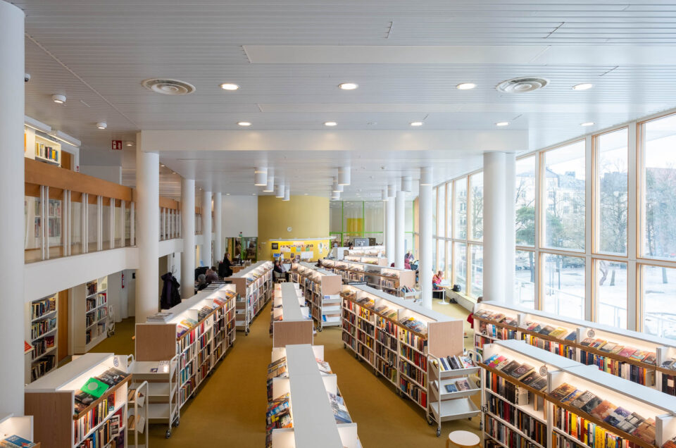 Large, modern library interior with tall white columns, rows of bookshelves filled with books, high ceilings, and large windows letting in natural light. Several people are sitting and reading or browsing books.