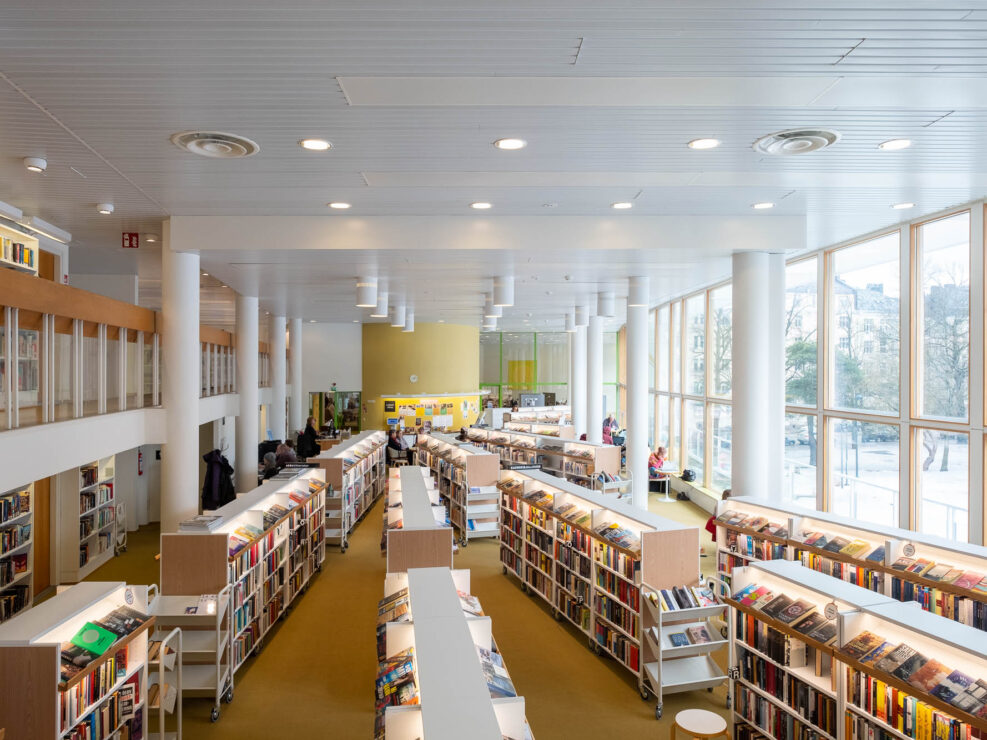 Large, modern library interior with tall white columns, rows of bookshelves filled with books, high ceilings, and large windows letting in natural light. Several people are sitting and reading or browsing books.