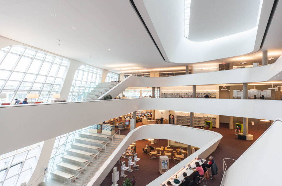 Interior of a contemporary library with curved white balconies, large windows allowing natural light, multiple floors with bookshelves, and people reading or working at tables on the lower level.