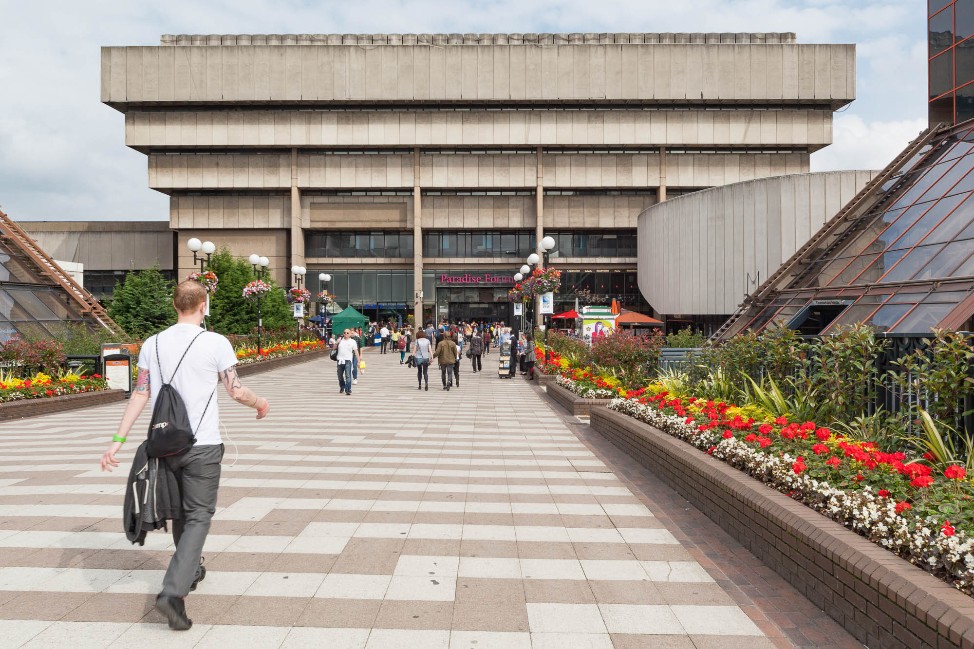 A man walks toward a large concrete building designed as an inverted pyramid. The path is lined on both sides with flowers in brick planters.