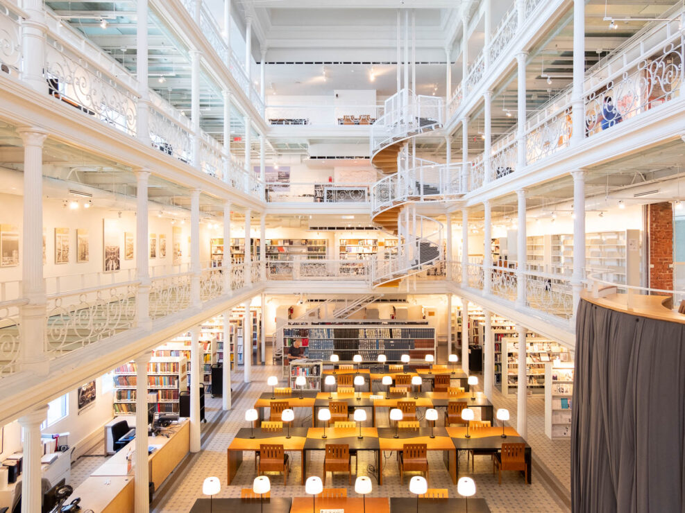 Interior of a library reading room surrounded by three floors of wrap-around mezzanines supported by Victorian-era iron columns and railings.