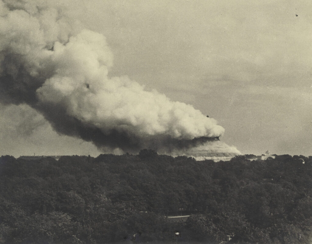 Black and white image of a burning building with a large plume of smoke rising over a densely wooded area.
