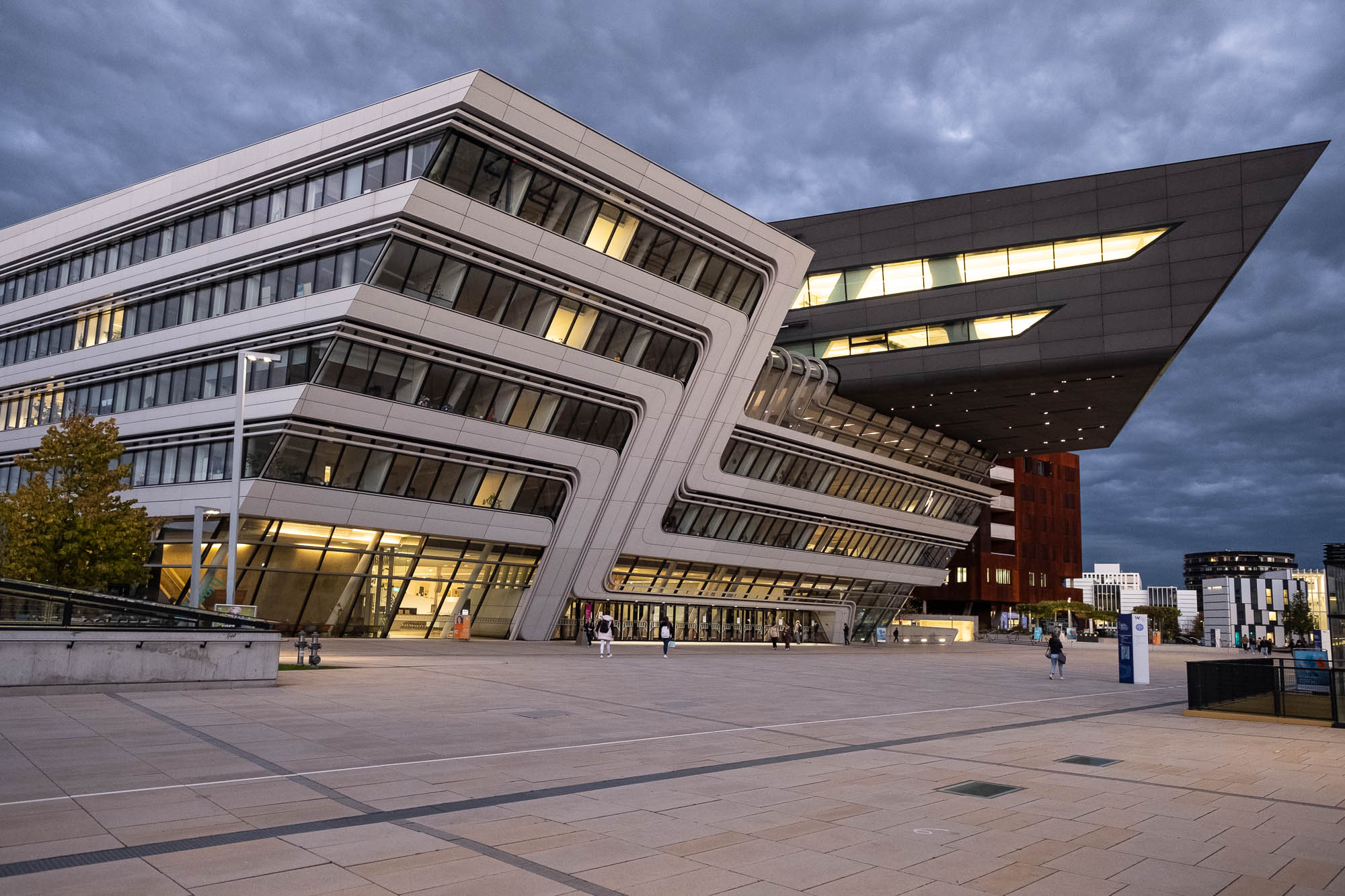 Futuristic architectural building with angular design, featuring illuminated windows and a dramatic overhang. The structure is set against a cloudy sky, with a few people walking on the wide, empty plaza in the foreground.