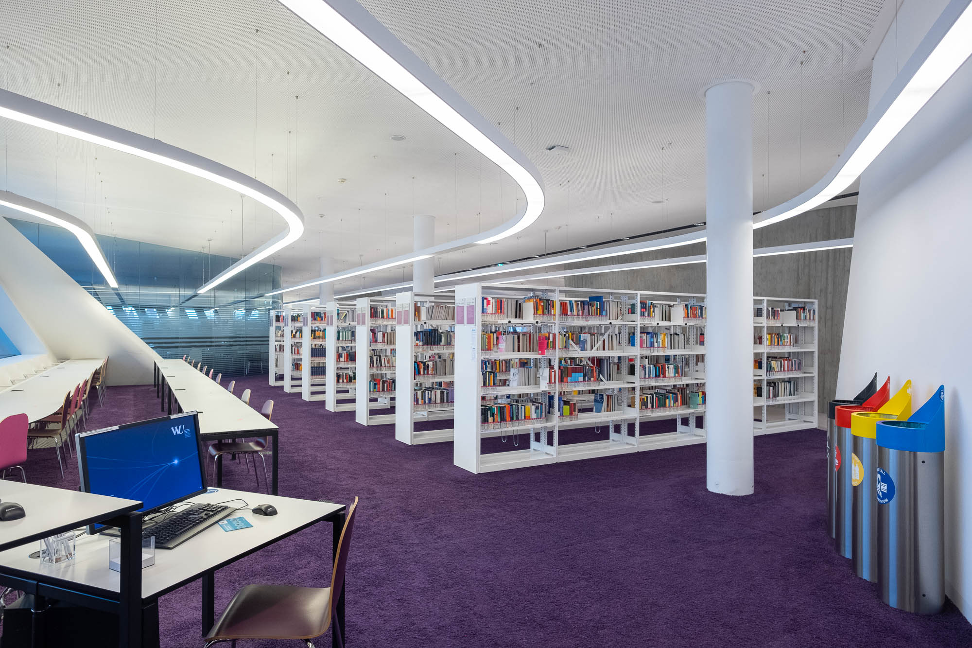 Futuristic library interior with a purple carpet and white bookshelves. There is a desk with a computer in the foreground and trash cans to the right.