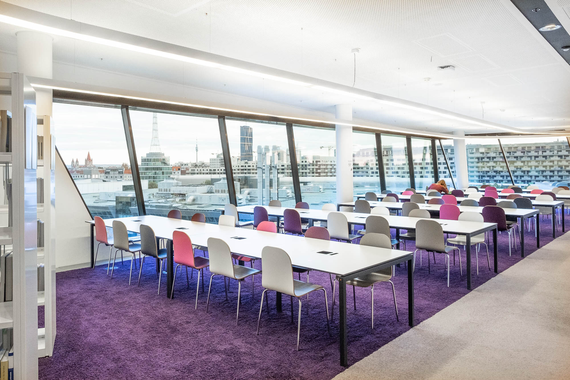 Contemporary library reading room with long white tables and colourful chairs, featuring floor-to-ceiling windows overlooking a cityscape. The floor is carpeted in purple.
