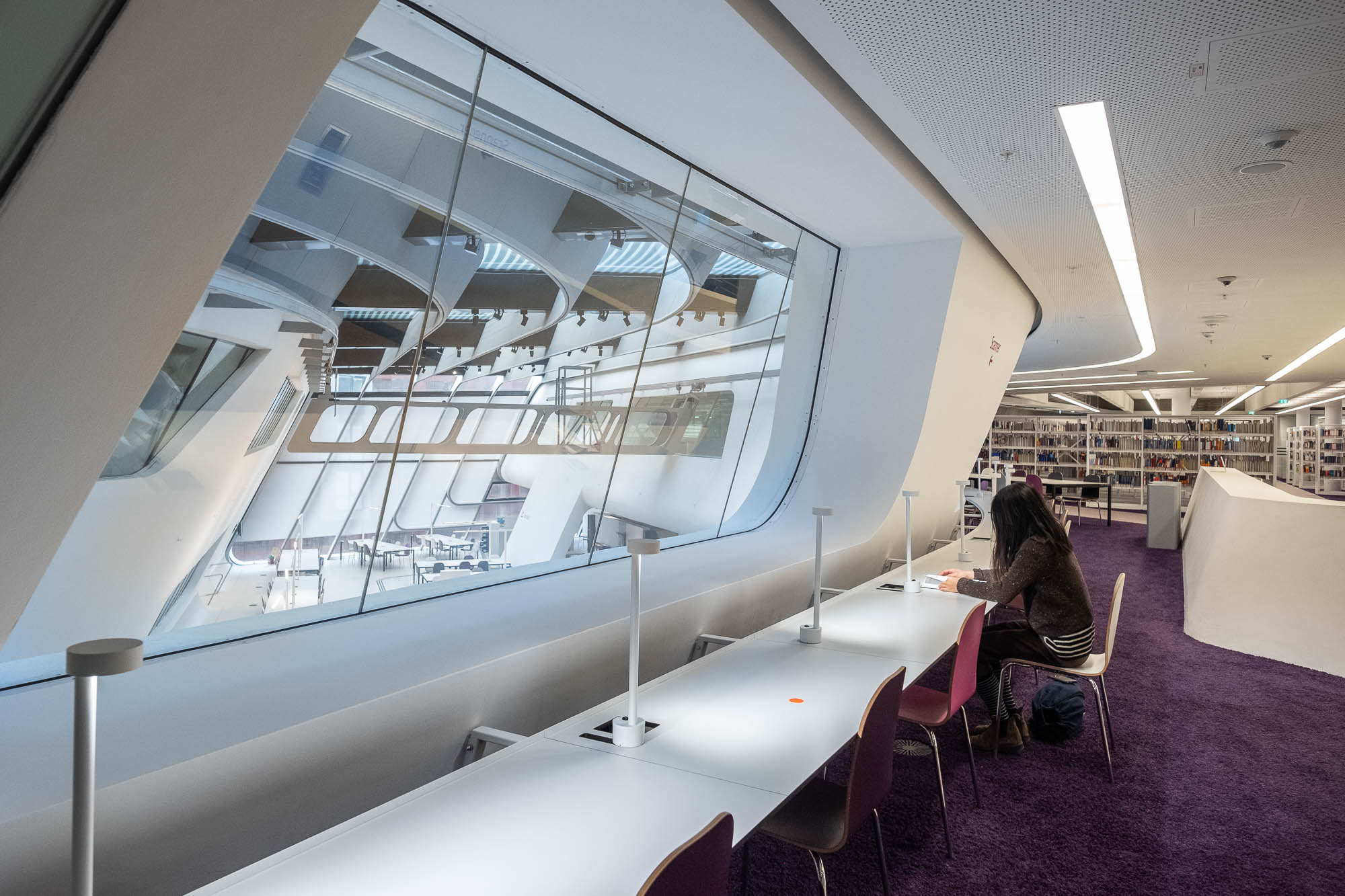Futuristic library interior with large windows and sleek white bookshelves. A large window in the foreground opens to an atrium with tables. A long table sits in front of the window, at which a person with long hair sits reading.