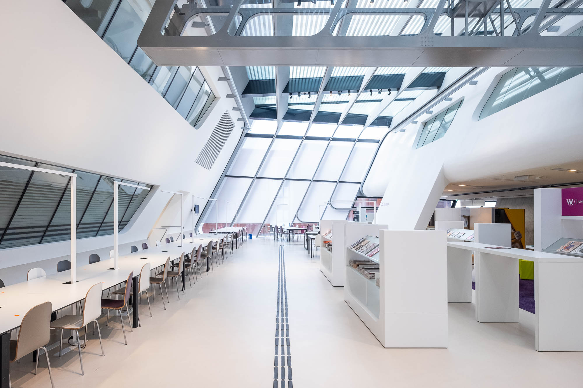 Futuristic library interior, a large open area with slanted walls and white furniture. A metal catwalk is visible on the ceiling.