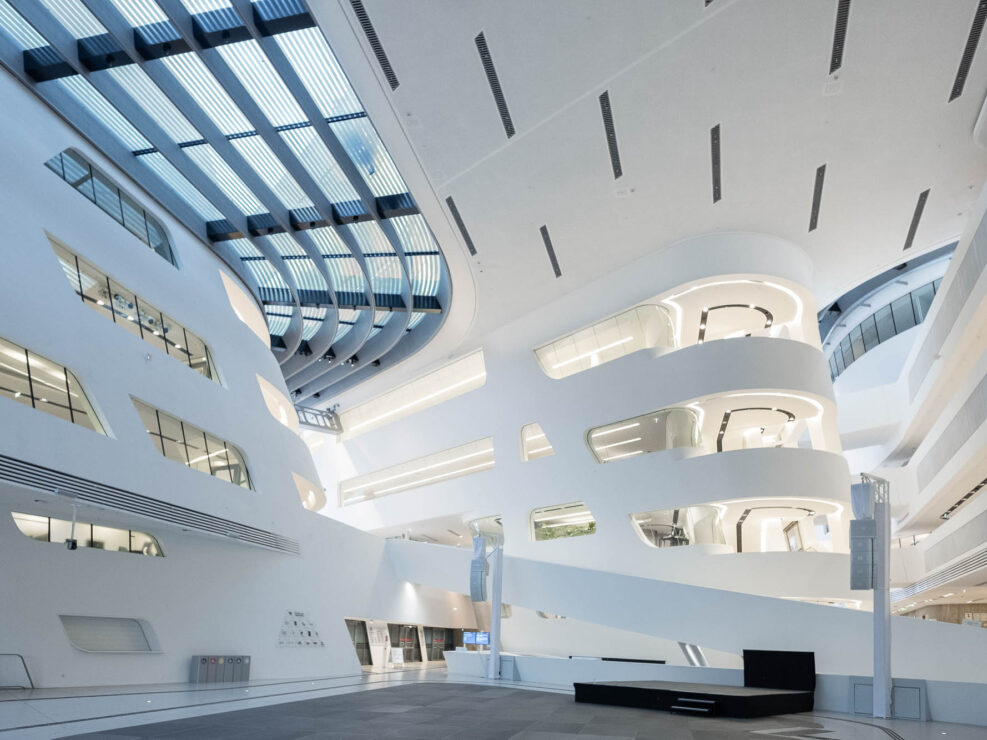 Futuristic architectural interior featuring curved white walls, large windows, and multiple levels. The ceiling has linear light fixtures and skylights.