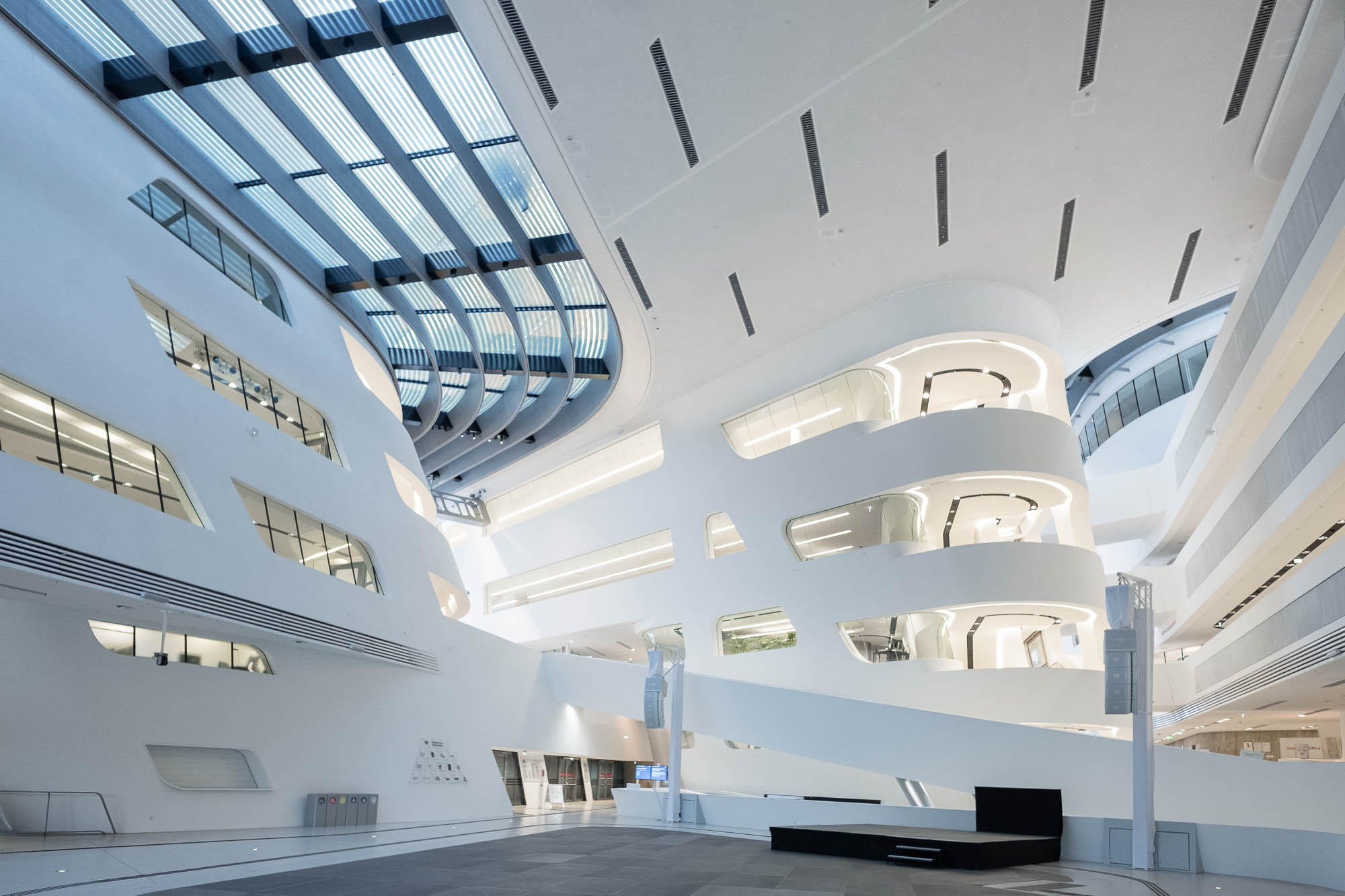 Futuristic architectural interior featuring curved white walls, large windows, and multiple levels. The ceiling has linear light fixtures and skylights.