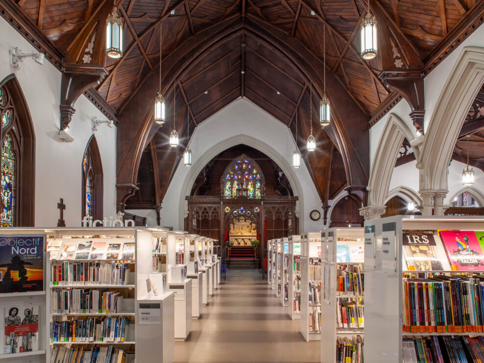 The image shows an interior of a library located in a former church. It features rows of bookshelves and high, arched wooden ceilings. Stained glass windows and ornate columns are visible, along with tables and chairs for reading.