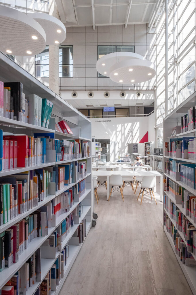 View of the main atrium of the Burgundy School of Business Learning Centre in Dijon, France. The image is taken from the ground level, with bookshelves on either side. The back of students working on long tables appear in the middle.