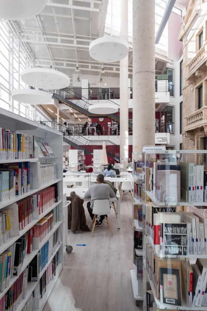 View of the main atrium of the Burgundy School of Business Learning Centre in Dijon, France. The image is taken from the ground level, with bookshelves on either side. The back of students working on long tables appear in the middle. The back wall shows two stacked mezzanines connected by stairs.