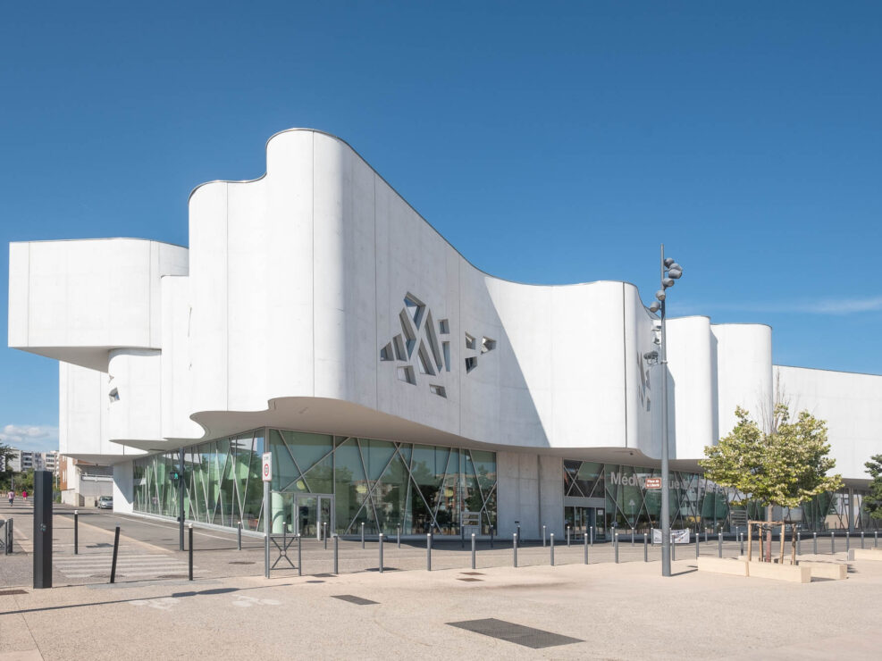 Exterior of a contemporary library. It has a wavy concrete facade with clusters of small irregular windows on a glass pedestal.