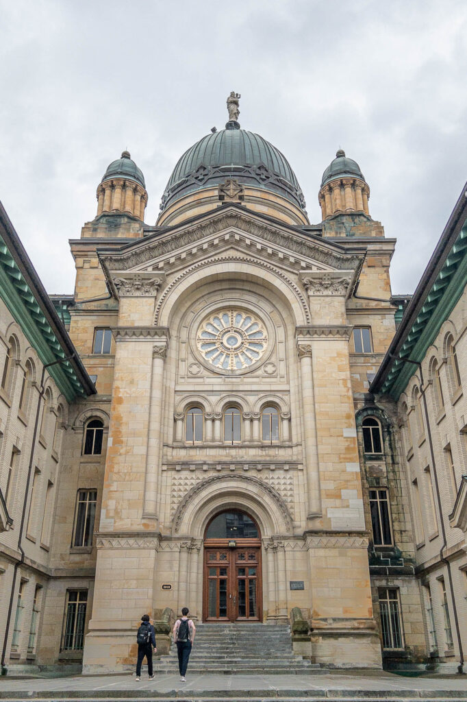 Exterior of a Beaux-Arts college in a converted convent, showing a tan stone and brick facade pierced by vaulted windows and a rosace and three cupolas on the roof.