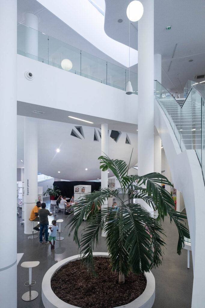 Interior of a contemporary library with white curving walls. The interior is mostly white, contrasted only with large green plants in the foreground.