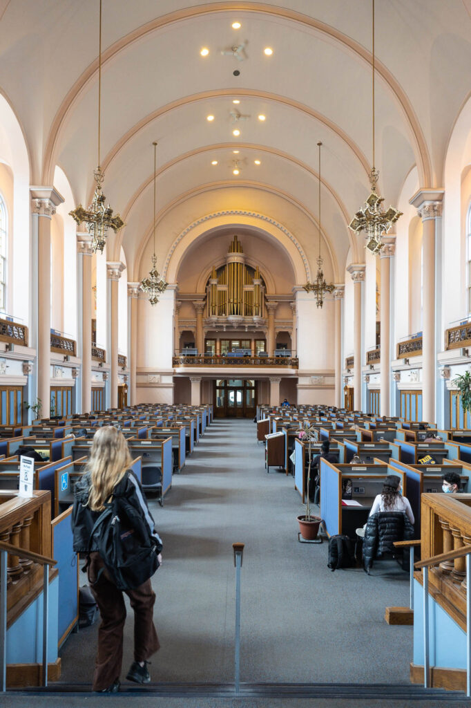 Interior of a library in a converted Neoclassical chapel. It is a pastel-coloured barrel-vaulted hall filled with small cubicles. An organ fills a gallery at the end of the room. Cross-shaped chandeliers hang from the ceiling.
