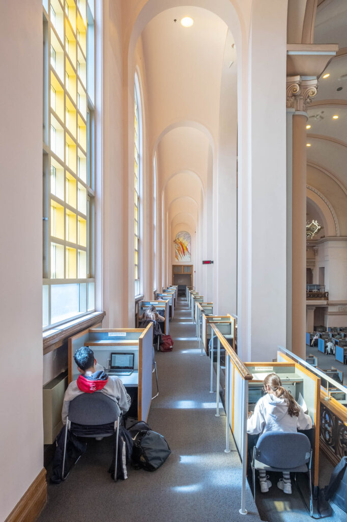 Interior of a library in a converted Neoclassical chapel. Pictured is a narrow vaulted side gallery, where cubicles are arranged on either side of a corridor. There are stained glass windows to the left. Students are working at the cubicles.