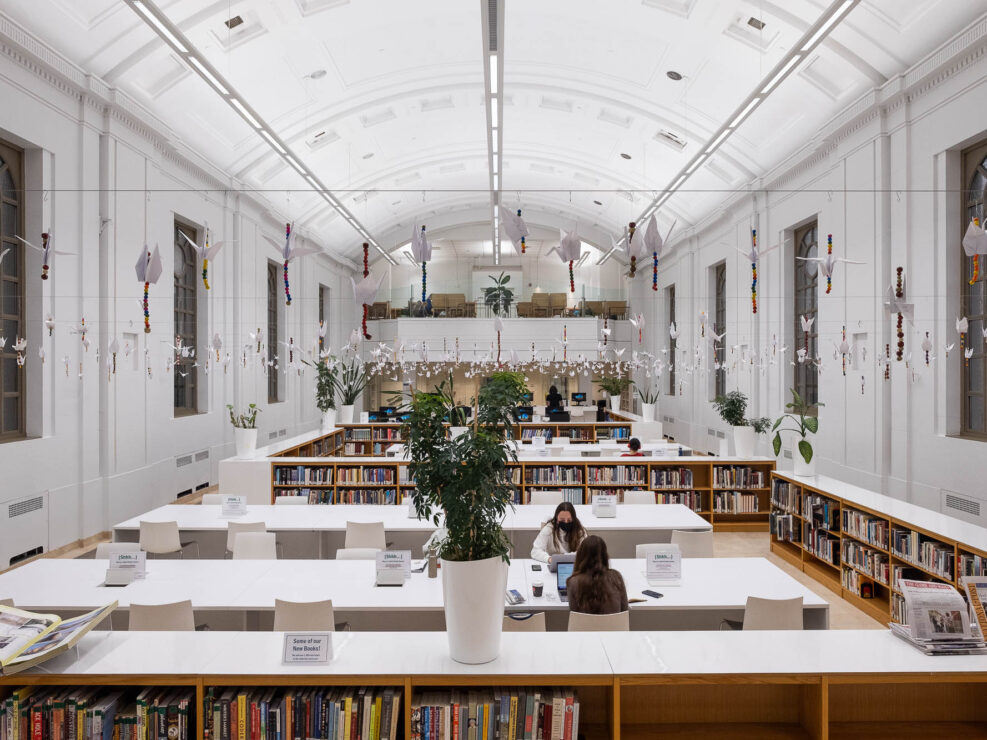 Interior of a contemporary library. It is a whitewashed rectangular room with a barrel-vault ceiling. The room is occupied by low bookshelves and long tables, where a few students are working. Large houseplants are set on the shelves. Paper cranes hang from cables strung across the room.