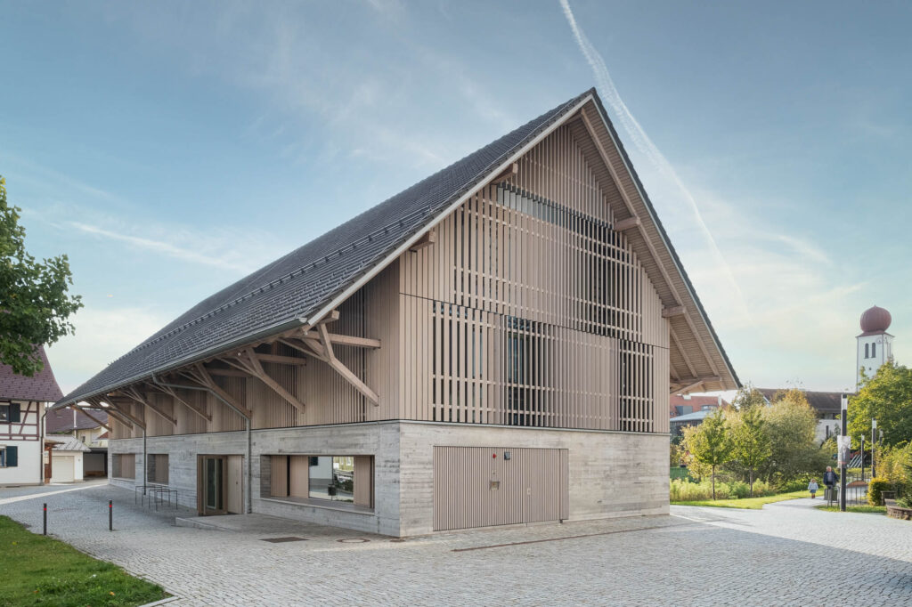 Exterior view of the library. It shows a barn-like structure with a concrete base over which a contemporary timber facade is built. The village church can be seen in the background.