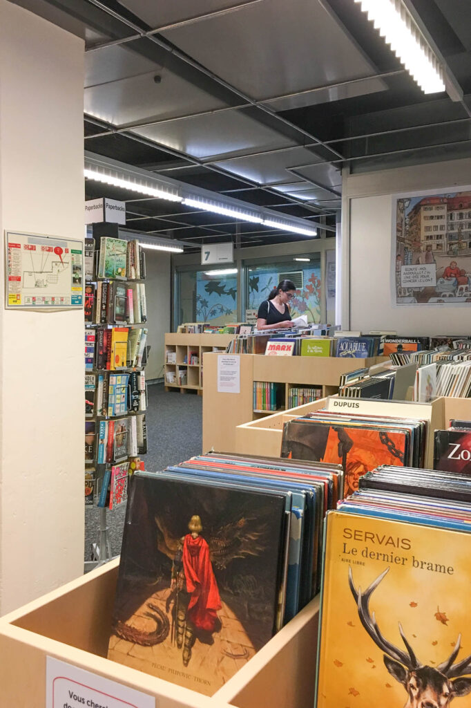 Interior view of a library, showing boxes of graphic novels. At the back, a woman is browsing the books.