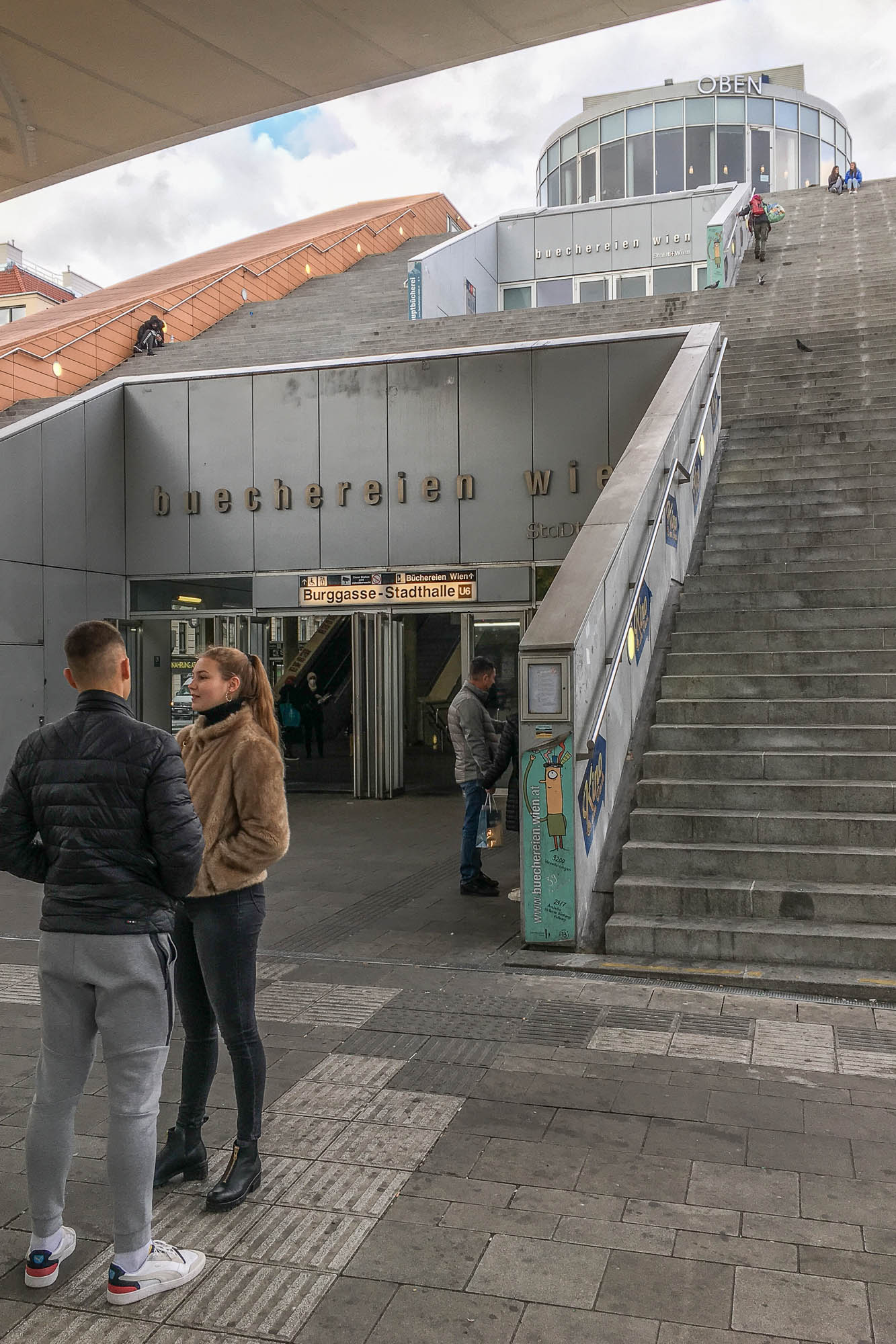 Exterior view of a large set of stairs, split in half to provide access to a subway station marked Burggasse-Stadthalle. On top of the subway entrance is another entrance to the library marked Buechereien Wien. A couple stands in the foreground.
