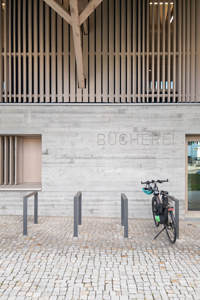 Exterior shot of the library and close-up on the facade; the lower half of the facade is concrete and the top half is covered in vertical timber slats. The word "Bücherei" (library) is stamped in the concrete. A bike rack is installed in front of the library, and a bike is attached there.