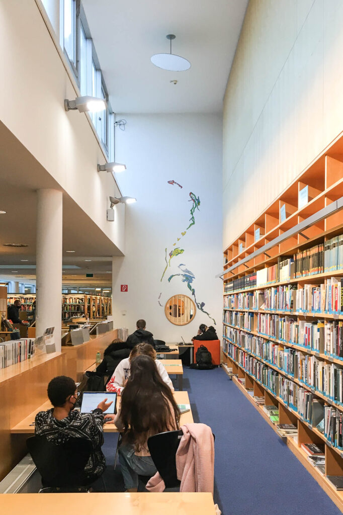 Interior view of a public library. A large bookshelf lines the wall to the right. In front, a row of tables is occupied by people talking and working on computers.