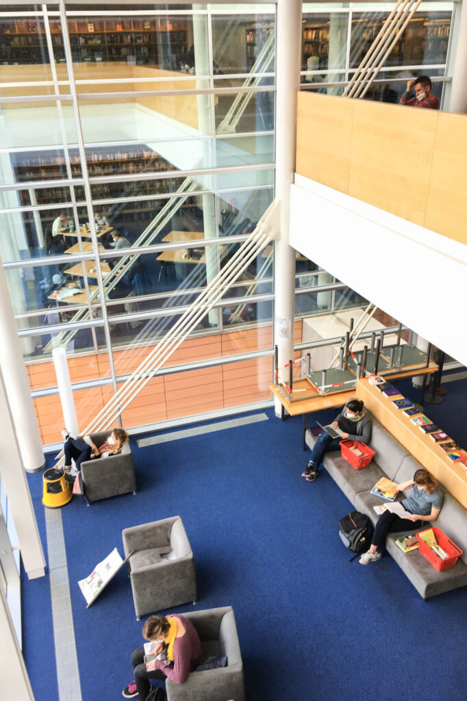Interior view of a contemporary library. It's a double-height glass-enclosed room with blue carpeting and patrons wearing face masks sitting on couches, reading. The image is taken from the upper floor, looking down inside the room.
