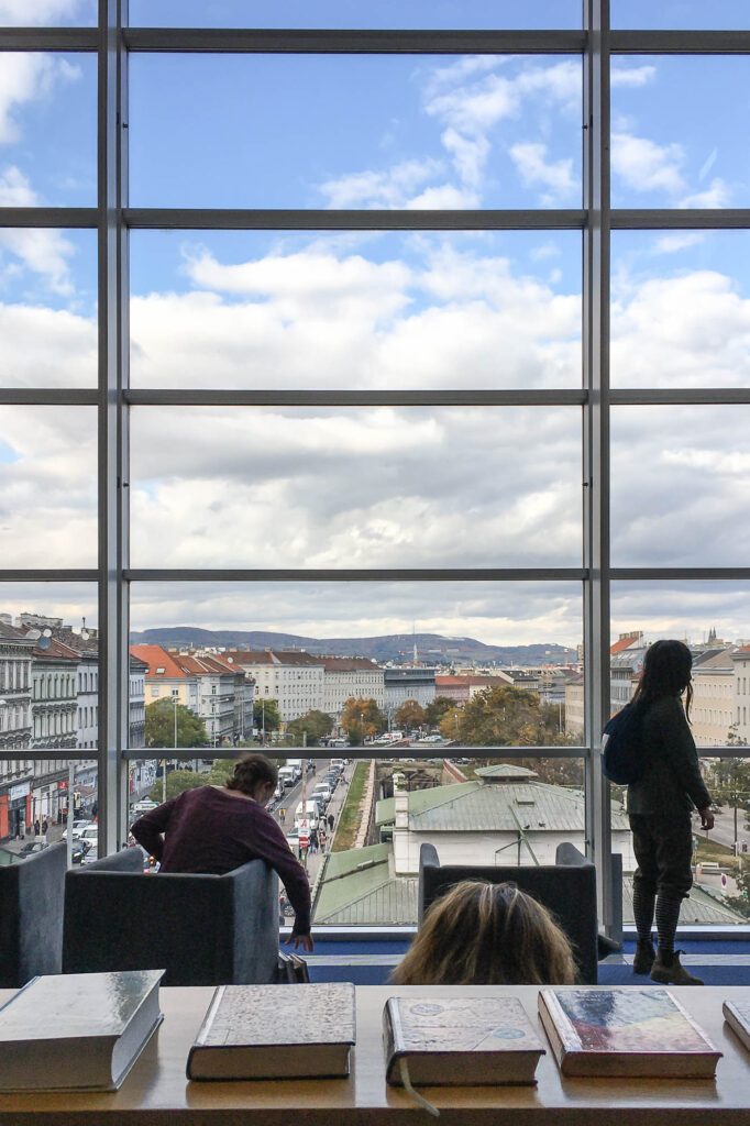Interior view of a library, most of the image is occupied by a large window through which the city of Vienna can be seen. Silhouetted library users stand in front of the window.