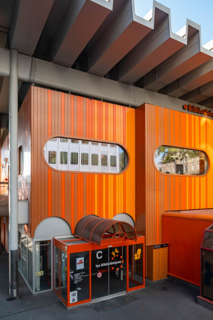 Exterior view of a 1970s pavilion with crenellated roof. The facade is made of vertical stripes of varying shades of orange and pierced with oval windows.