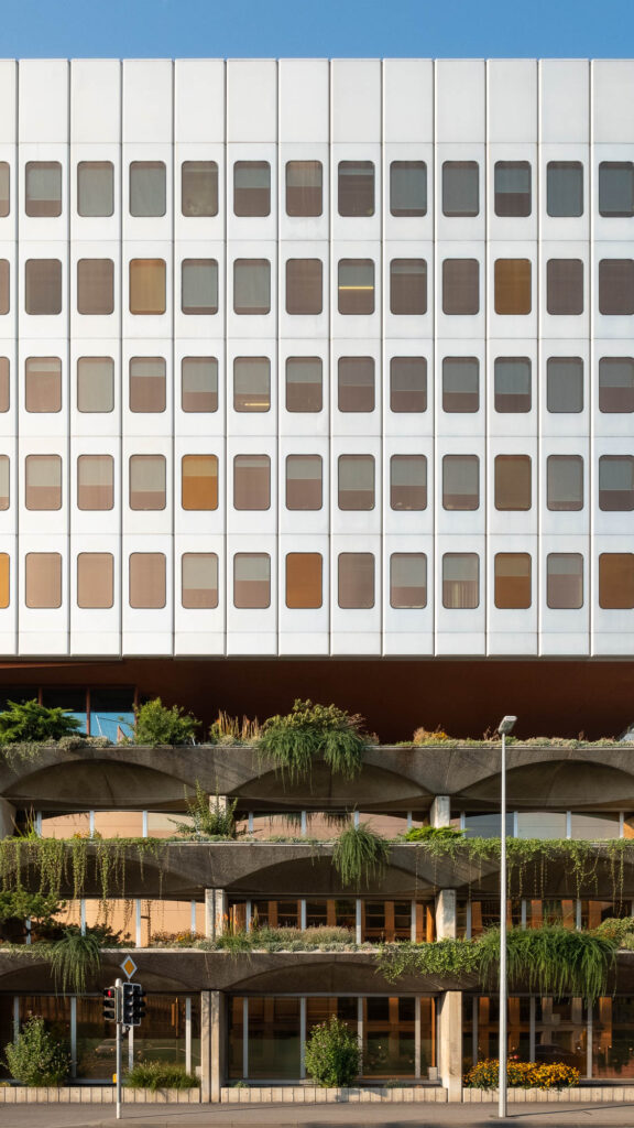 Exterior of a 1970s office building. The lower part has raw concrete planters with wide arches, overflowing with vegetation. On top is a five-level high-rise with a facade of light-coloured metal elements surrounding rectangular windows with rounded corners. Windows are tinted orange.