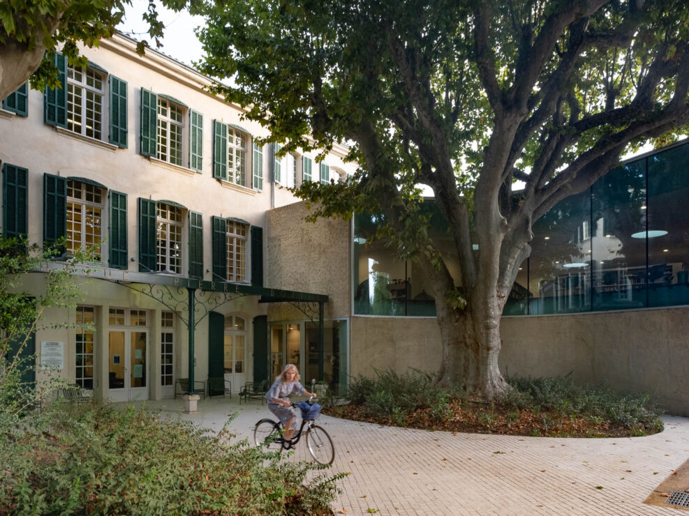 Exterior of an old house converted to a library. The original building to the left is a three-story house with green shutters and a wrought iron pergola. The modern wing of the library to the right has a curved wall and window to accommodate a large mature tree growing in front of the house. A person on a bicycle moves through the scene.