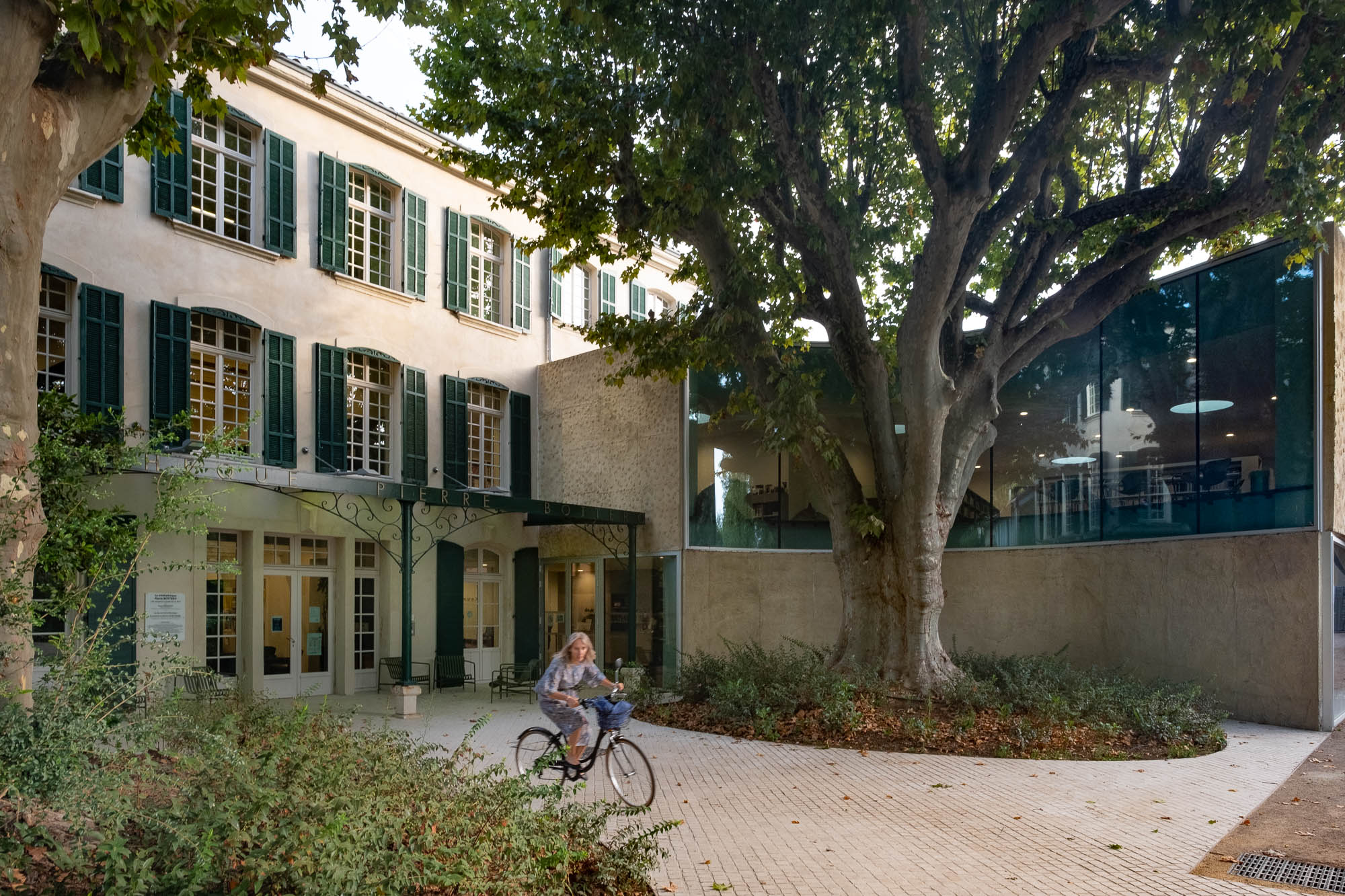 Exterior of an old house converted to a library. The original building to the left is a three-story house with green shutters and a wrought iron pergola. The modern wing of the library to the right has a curved wall and window to accommodate a large mature tree growing in front of the house. A person on a bicycle moves through the scene.