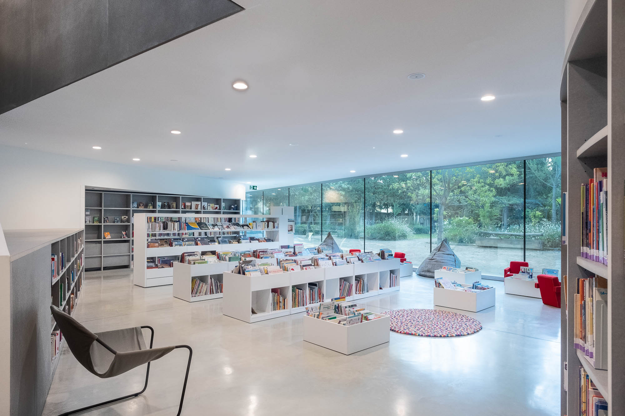 Interior of a modern library. A large window is seen at the back, opening to a garden. In the room are low bookshelves, book boxes, bean bags and a colourful rug.