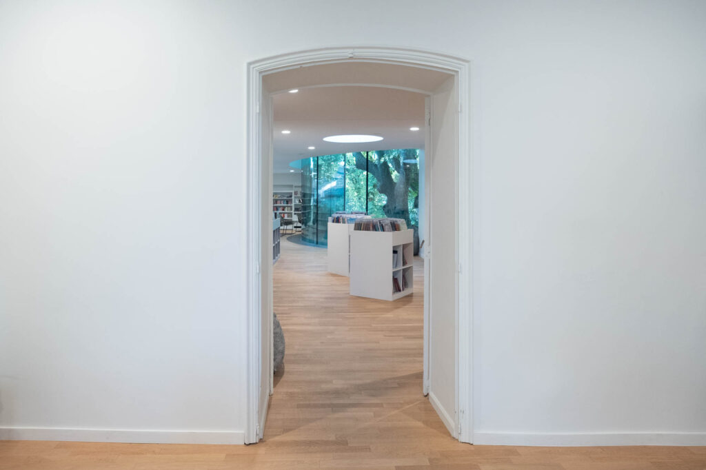 Interior of a library, doorway from an older part of the building towards a modern wing, through which a curved window can be seen.