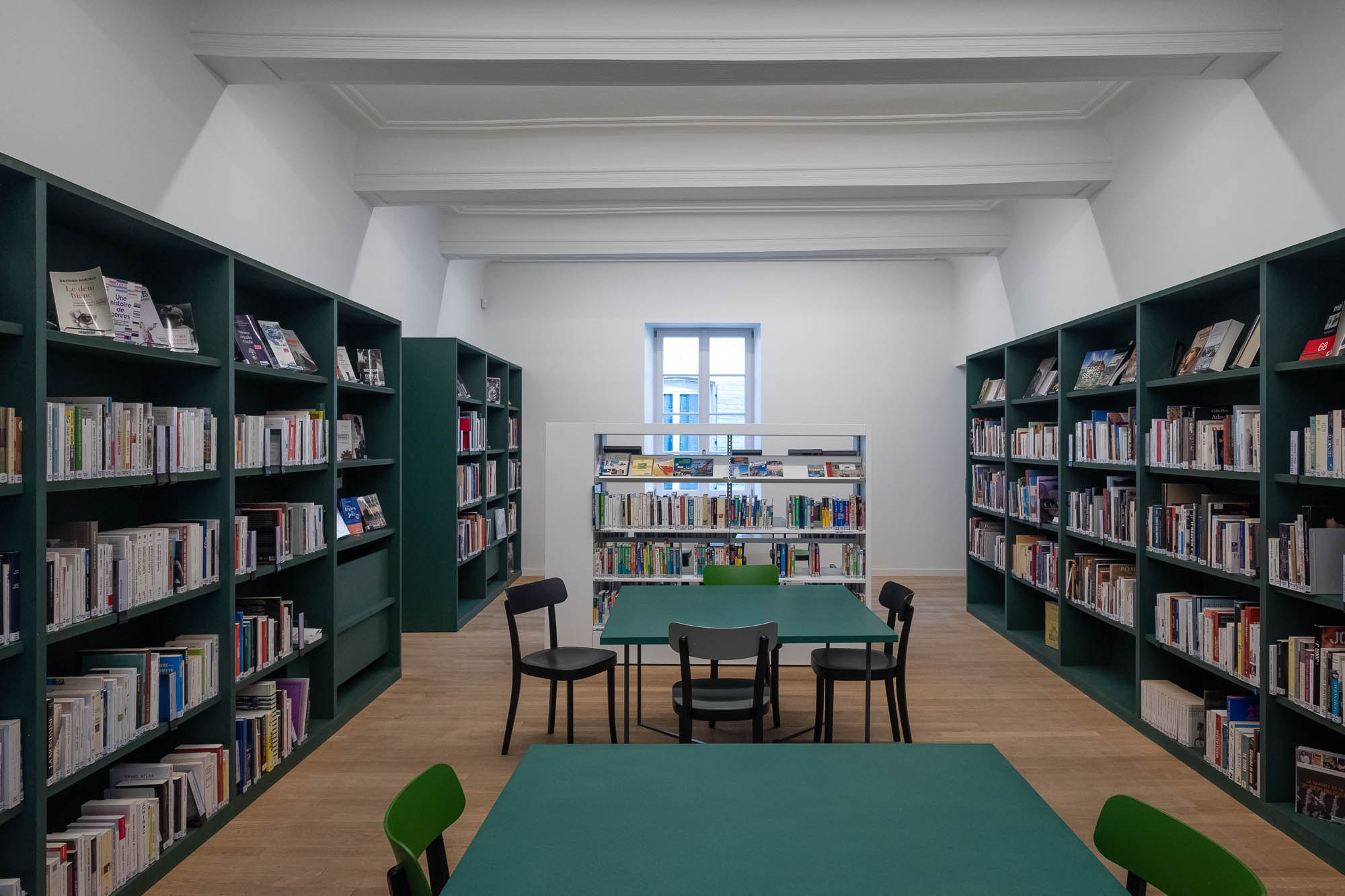 Interior of an old house converted to a library. Dark green bookshelves and tables contrast against white painted walls and ceilings, where beams are visible.