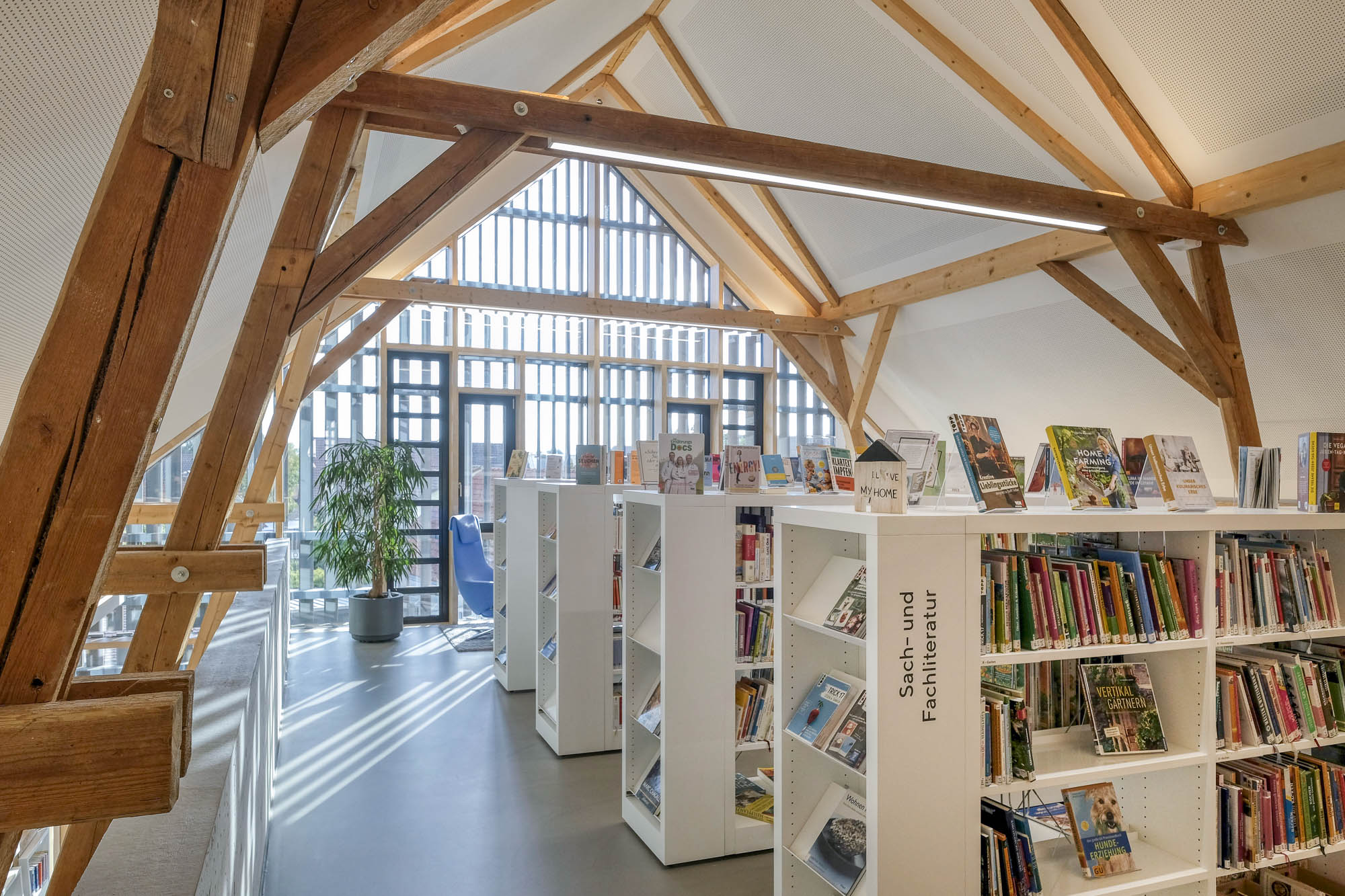 Library interior showing a row of white bookshelves filled with books. A large window opens at the back. The roof is supported by antique wooden beams.