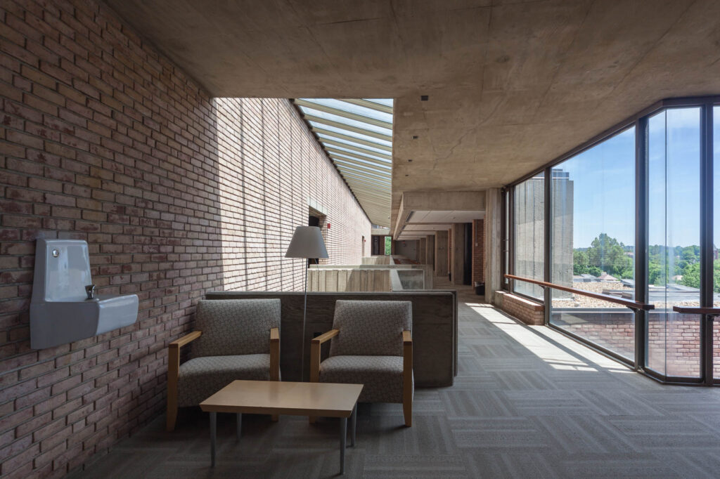 Interior view of a brick and concrete brutalist library, towards a corridor and light well separating the bricked book tower to the left and exterior windows to the right. A couple of comfy chairs, low table and floor lamp are visible in the foreground.