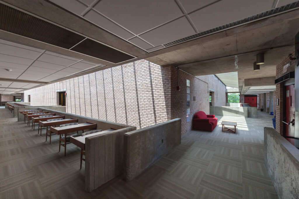 Interior view of a brick and concrete brutalist library. It is an angled shot showing a corner of the central brick core of the library in the middle surrounded by light wells and corridors. To the left, a series of cantilevered work carrels is set against the parapet.