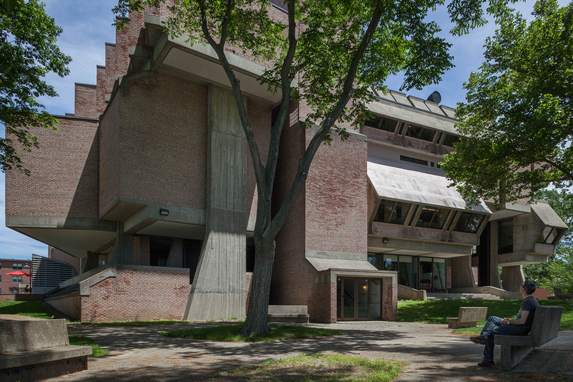 Exterior view of a brick and concrete brutalist library, showing a variety of shapes and forms. There are trees in front of the library. A person sits on a concrete bench, looking up at the library.