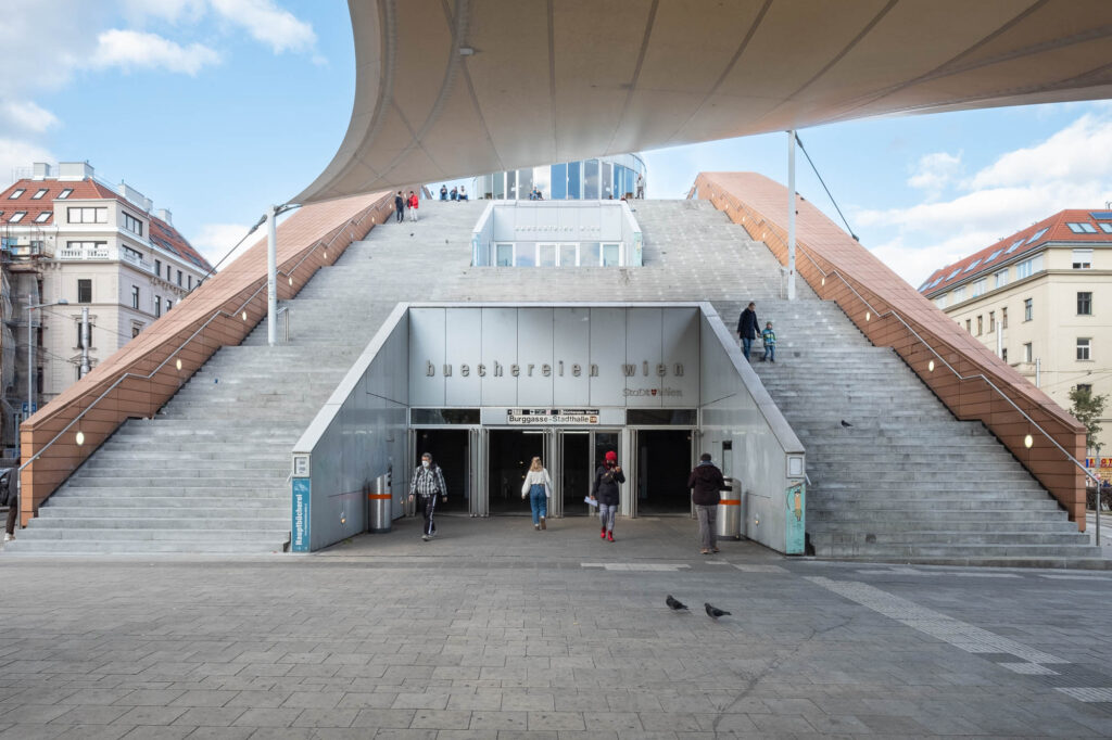 Exterior view of a large set of stairs, split in half to provide access to a subway station marked Burggasse-Stadthalle. On top of the subway entrance is another entrance to the library marked Buechereien Wien. People are entering and exiting the station and climbing down the stairs.