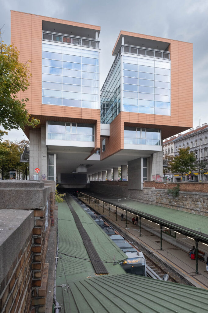 Exterior view of a contemporary building on concrete stilts straddling a light rail station recessed into the ground.