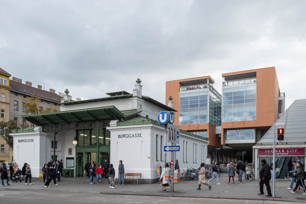 Exterior view of a Jugendstil subway stop marked Burggasse and surrounded by commuters. Behind the station is a contemporary glass and orange cladding building composed of two symmetrical shapes separated by a narrow space.