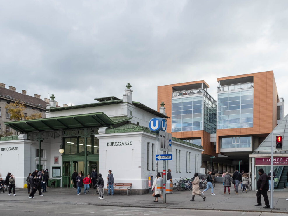 Exterior view of a Jugendstil subway stop marked Burggasse and surrounded by commuters. Behind the station is a contemporary glass and orange cladding building composed of two symmetrical shapes separated by a narrow space.