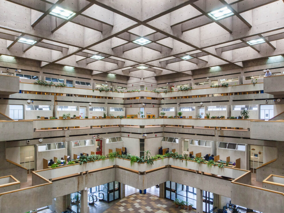 Interior of a brutalist library atrium, with cascading terraces lined with planters, in which lush green interior plants grow. Students are working at cubicles. Boxy staircases lead from one level to another. A person stands on top of the topmost staircase and looks at the atrium below.