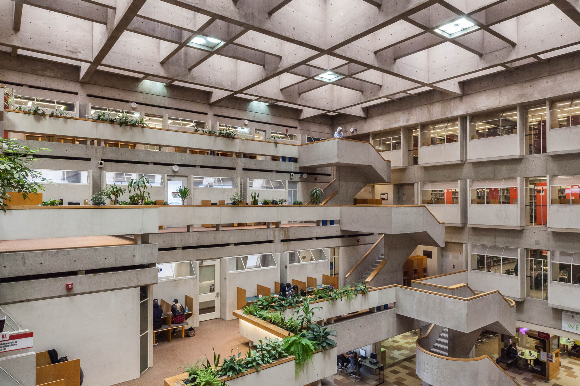 Interior of a brutalist library atrium, with cascading terraces lined with planters, in which lush green interior plants grow. Students are working at cubicles. Boxy staircases lead from one level to another. A person stands on top of the topmost staircase and looks at the atrium below.