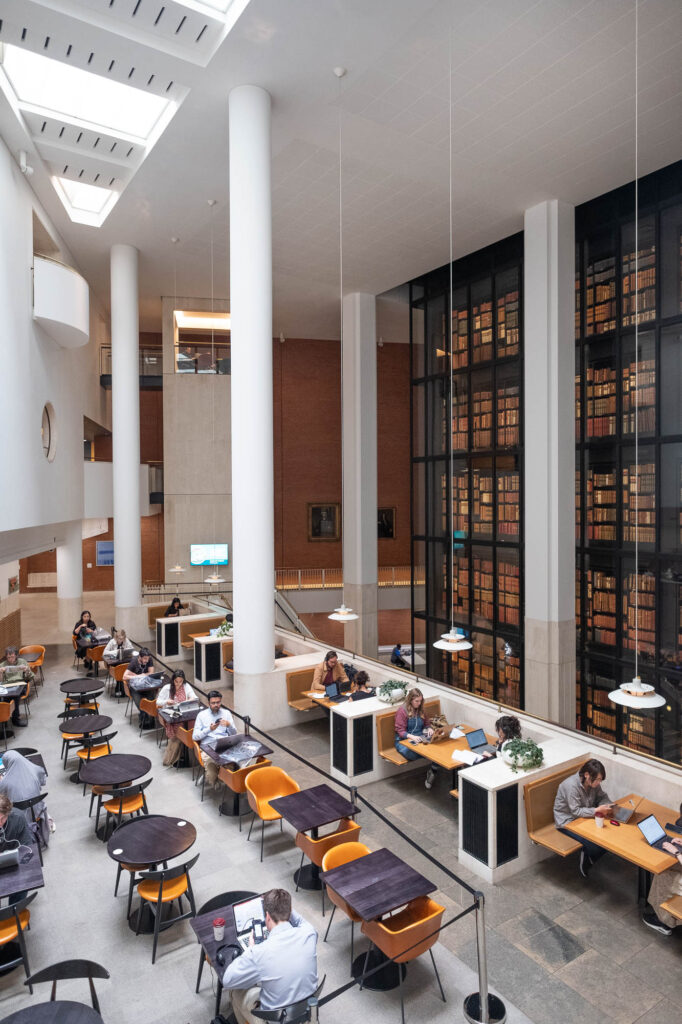 Interior view of a modern library. To the right and in the back is of a dark steel and glass facade through which rows of rare books are seen, illuminated in a warm glow. The foreground is occupied by a seating area with small cafe tables with people working on them.