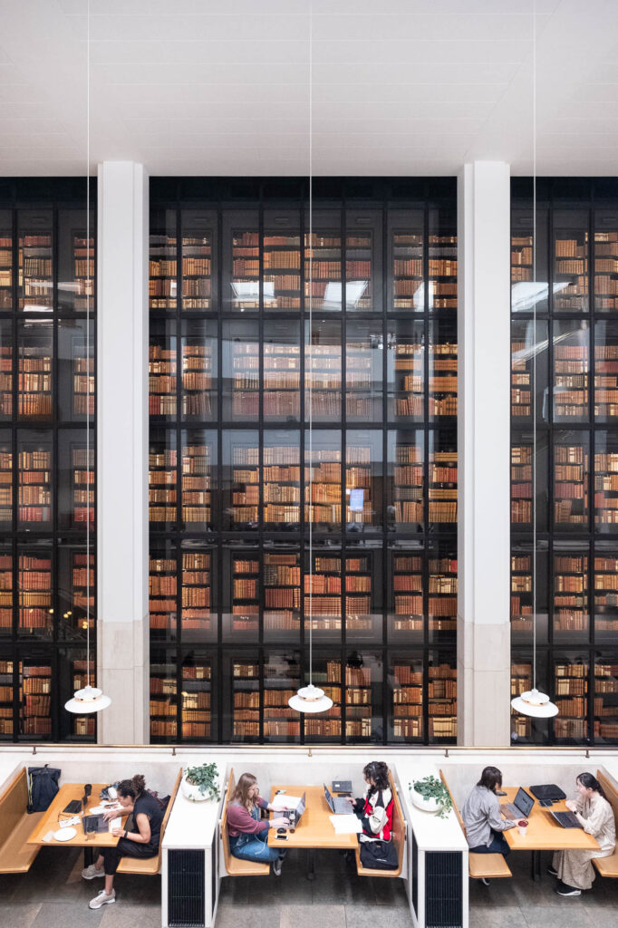 Interior view of a modern library. The background is of a dark steel and glass facade through which rows of rare books are seen, illuminated in a warm glow. In the front is a row of work booths with people working on laptops.