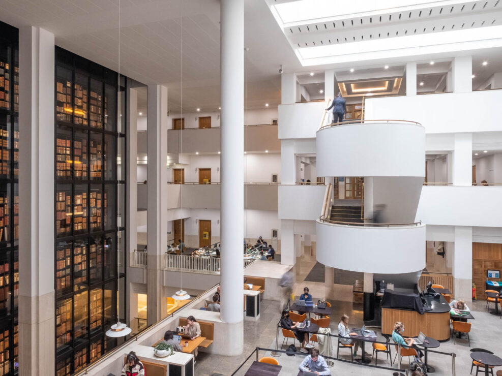 Interior view of a modern library. On the left is a large black glass box filled with rare books that glow in a warm light. A staircase with people moving on it is seen in the middle. In the front is a seating area with small cafe-like tables and people working on laptops.